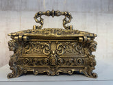 An ornate antique-style brass trinket box with a decorative handle, featuring intricate scrollwork and sculpted faces on its sides, sitting on a white surface.